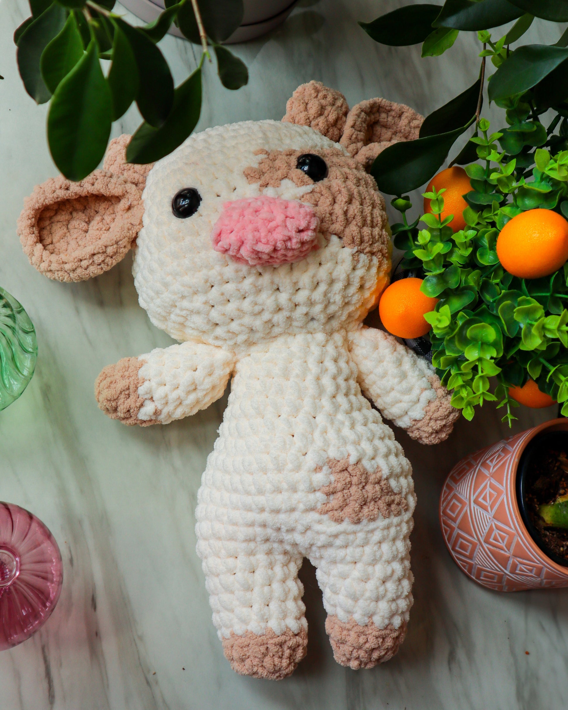 Crocheted animal toy with pink and brown accents on a light surface with plants and fruits. Crochet cow.