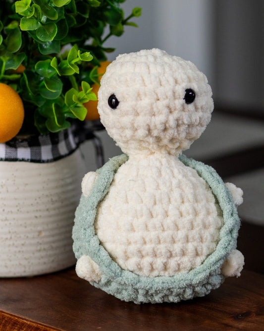 Crocheted turtle toy on a wooden surface with a plant in the background background